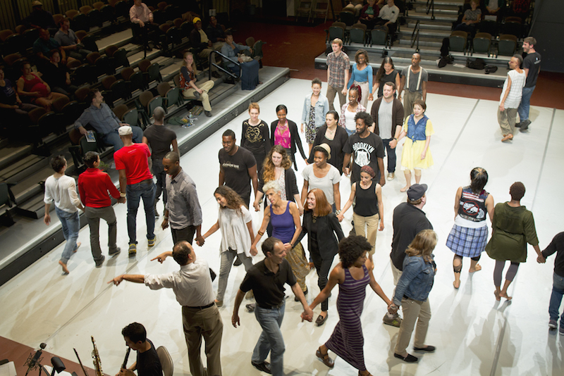 A large group of people walk throughout a theater space while an audience in the round witnesses. Some hold hands.